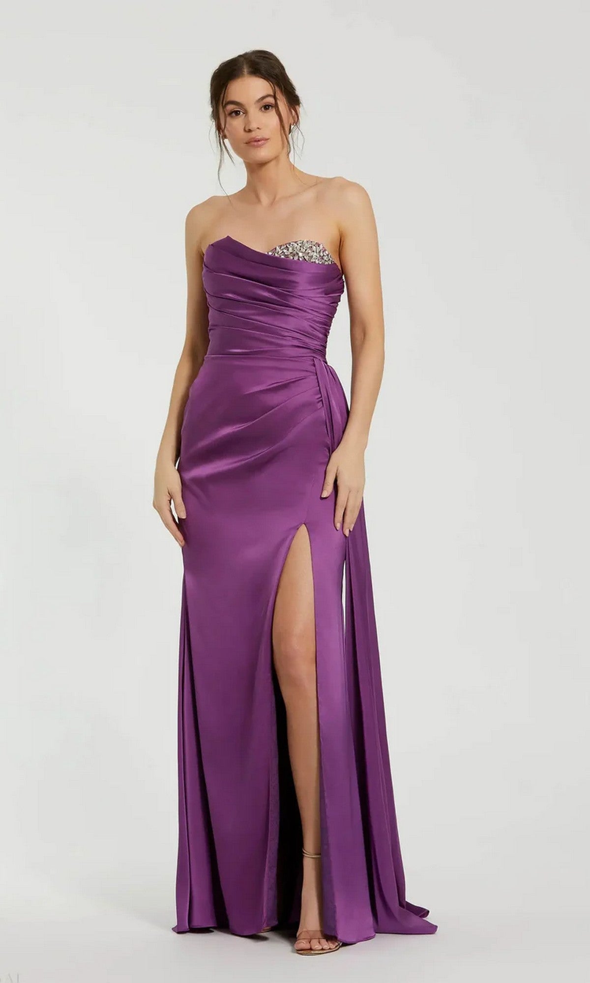 A woman wears the Mac Duggal 2211 Side-Drape Strapless Prom Gown in purple satin with a pleated bodice, side slit, and embellished details, standing against a plain background.